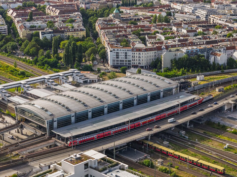 Aerial view of modern Ostkreut train station with red passenger trains arriving at the platforms, surrounded by urban residential buildings and railway tracks in Friedrichshain, Berlin, Germany.