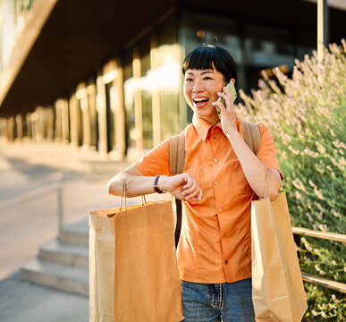 Portrait of a happy asian woman holding shopping bags, using a smartphone mobile phone and walking down the street