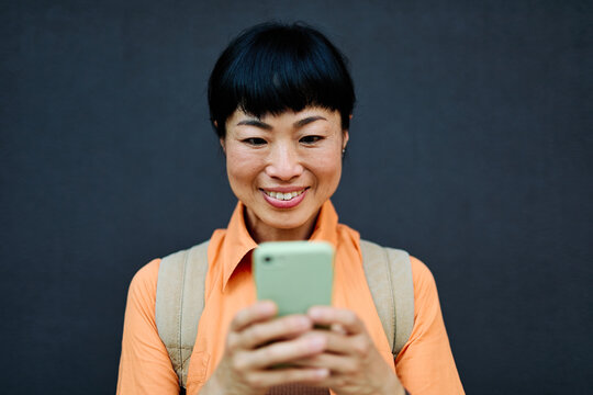 Portrait of a happy asian woman posing, using a smartphone phone or taking a selfie photo outdoors in front of a modern urban background
