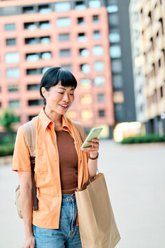 Portrait of a happy asian woman holding shopping bags, using a smartphone mobile phone and walking down the street