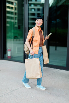Portrait of a happy asian woman holding shopping bags, using a smartphone mobile phone and walking down the street