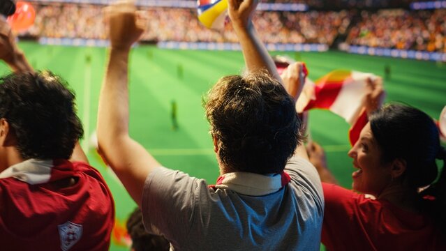 Soccer fans in red shirts react with excitement to goal scored during match, passionate supporters in stadium grandstands following game action on green pitch for football fan and match day concepts.