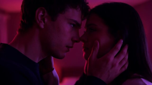 Passionate couple looking at each other before kissing in a purple neon light filled bedroom