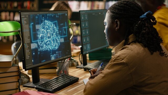 Black student working with AI brain display on desktop computer in library, studying automation in computer class with peers.