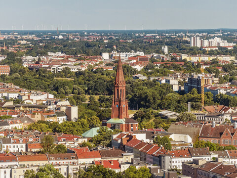 Aerial view of a prominent red-brick church tower rising above the surrounding urban landscape, rooftops, and lush greenery in Berlin, Berlin, Germany.