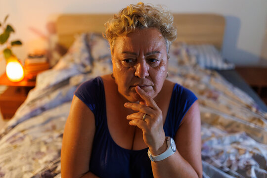 Depressed overweight older woman sitting lonely on bed during dark evening.