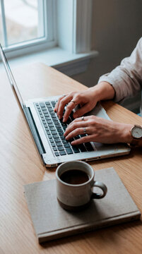 Woman typing on laptop with coffee mug on notebook at bright wooden desk