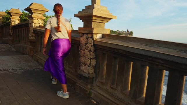 Young Woman in Traditional Purple Sarong Looking Toward Coastal Temple Cliffs at Pura Luhur Uluwatu During Golden Hour
