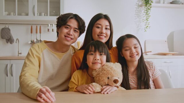 Smiling Happy Asian Family With Two Kids Sitting At Table In Bright Kitchen, Enjoying Time Together, Holding Teddy Bear, Creating Warm Family Memories, Home Lifestyle Concept.