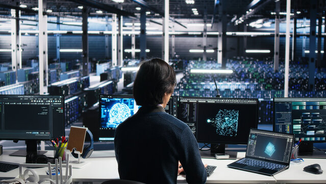 Computing facility system administrator managing AI systems for uptime and efficiency, while IT staff member working with AI technology on computer and notebook in computing hub.