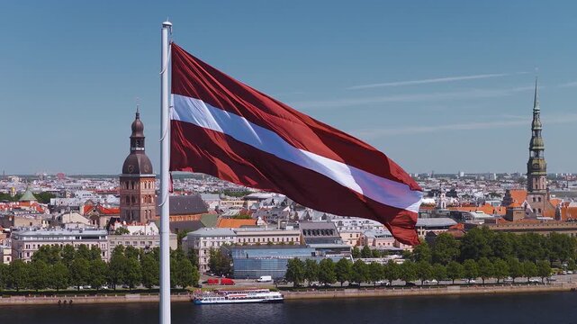 Aerial daytime view over the Daugava River in Riga, Latvia. A Latvian flag waves, a cruise boat moves, Riga Cathedral dome and St Peters Church spire rise among rooftops.