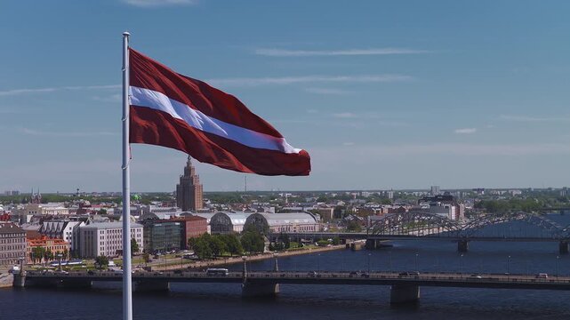 Aerial view shows Latvian flag billow above the Daugava River, central station arches, steel railway bridges, and Latvian Academy of Sciences in bright midday light.