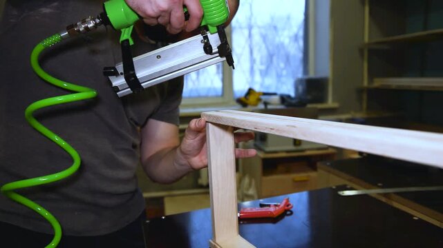 Master uses a pneumatic hammer to fasten the wooden planks, work with pneumatic tool, production of furniture and interior items, carpentry in the workshop.