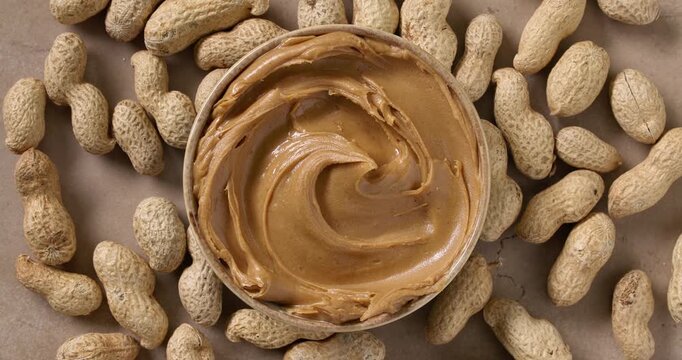 Smooth creamy peanut butter in a rotating bowl with a swirl on top, peanuts in the background. Slow motion, top view