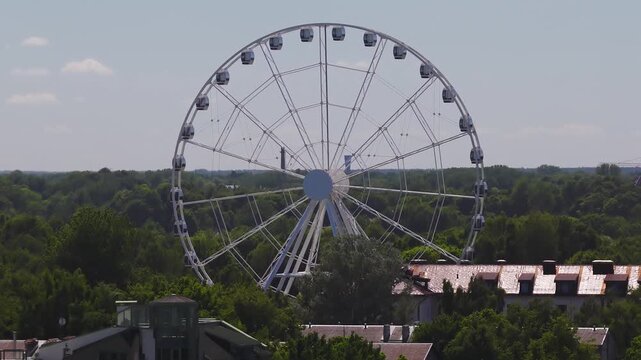 Aerial daytime view in Riga, Latvia shows a white Ferris wheel rising over tree canopy and red tiled roofs, its spokes and gondolas slowly rotate under pale sky.