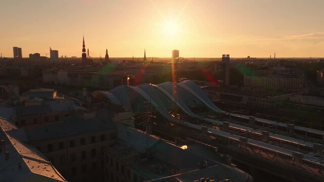 Aerial drone pan over Riga, Latvia, with Central Railway Station roof and platforms, Old Town spires of St. Peter's and Dome Cathedral, and distant Vansu Bridge in warm light.