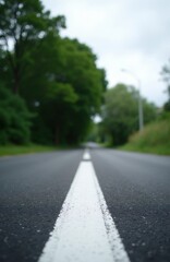 Fototapeta premium View from asphalt road surface with central white lane marking. Trees line road sides under cloudy sky. Perspective shot shows empty roadway vanishing into distance.