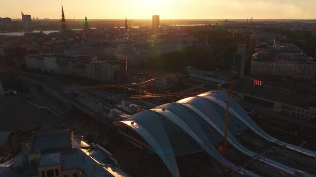 Aerial pan over Riga, Latvia shows Central Railway Station roof of Rail Baltic project, tower cranes, tracks, Old Town spires of St. Peter's and Dome Cathedral, Daugava, warm golden hour light.