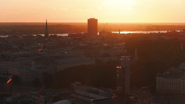 Aerial sunset scene in Riga, Latvia, with Old Town spires and clock tower, Daugava River, a tall riverside tower, warm light, slow pan, layered city to river depth.