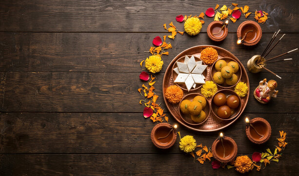 Indian Diwali festival celebration background with copy space for text, top view flat lay of traditional sweets mithai thali and burning diya lamps on dark rustic wooden table