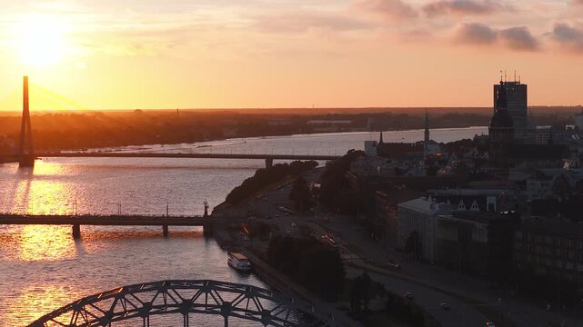 Aerial 4K sunset over the Daugava River in Riga, Latvia. Vansu Bridge sits left, Old Town spires and Riga Cathedral right. Cars move, a riverboat is moored.