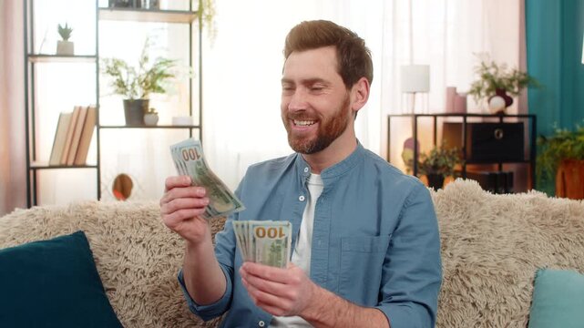 Young man at home holding a stack of dollar cash banknotes, counting money with excitement. Caucasian guy on sofa smiles, feeling happy about a big paycheck lottery win or unexpected financial success