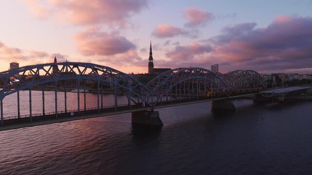 Aerial view shows arched steel railway bridge in Riga, Latvia, St. Peter's spire, yellow train crossing, boats moving, sunset sky, warm reflections on the Daugava.