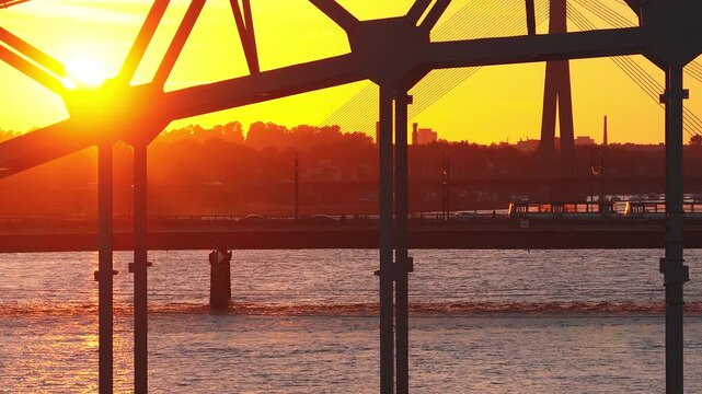 Aerial dusk view of the Daugava River in Riga, Latvia, as cars and a tram cross a steel bridge, Vansu Bridge pylons rise beyond, and warm reflections ripple.
