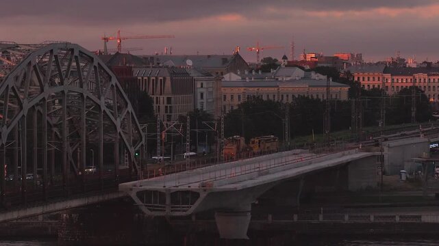 Aerial view of Riga, Latvia shows steel arch railway bridge and new concrete span, cranes, rail tracks, catenary masts, and moving cars at late evening under clouds.