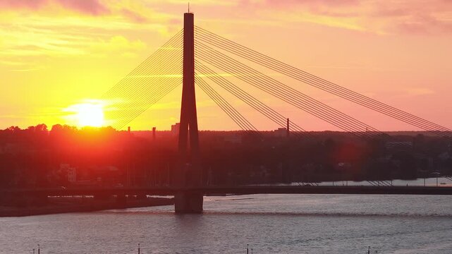 Aerial view of Riga, Latvia at sunset shows Vansu Bridge over the Daugava River, a tall pylon with parallel cables, rippling water, light traffic, and lens flare.