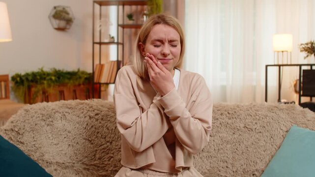 Young adult woman holds cheek in pain while sitting on home sofa, showing signs of strong toothache. Blonde girl frowns and shows discomfort, reacting emotionally to dental problem and sensitivity