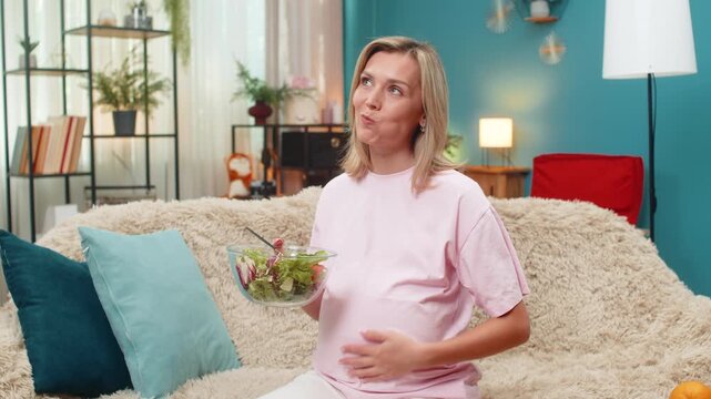 Pregnant middle-aged woman at home eats cabbage tomato salad enjoying vitamin nutrition aiding baby health. Caucasian girl on sofa smiles tasting veggies showing healthy pregnancy lifestyle joy mood.