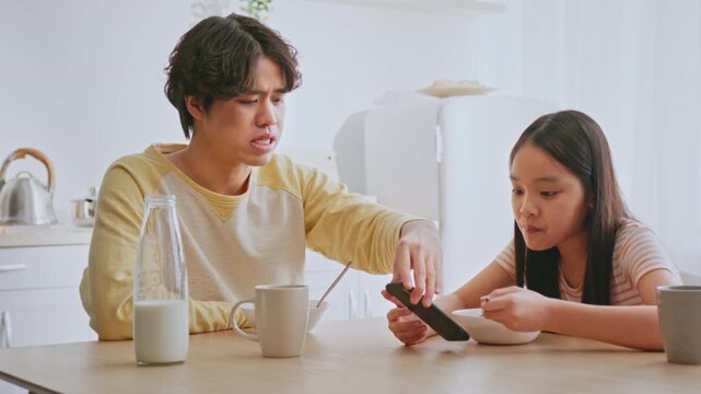 Concerned Young Asian Man Teaching Little Girl About Proper Phone Usage While Eating Breakfast At Home Kitchen Table, Sharing Parenting Tips. Emphasizing Responsible Technology Use And Family Time.