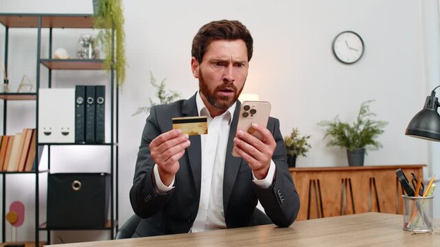 Middle-aged businessman at home office holds smartphone and credit card sees low balance sighs disappointed. Freelancer guy at table lowers gaze budget calmly after failed payment attempt discouraged.