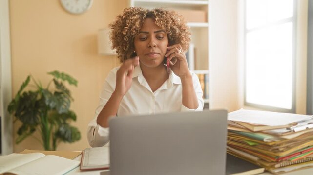 Woman holding phone to ear and typing on laptop with stacked folders and open notebook on desk in building; focused productivity.