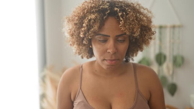 Woman in bra with bare shoulders looking at camera in a studio, curly hair framing face, soft natural light and a macrame plant hanger visible; vulnerability reflection.