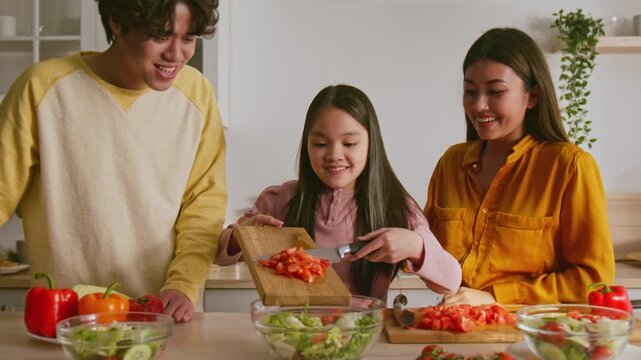 Smiling Asian Girl Slicing Fresh Tomatoes With Chef Knife While Family Assists In Kitchen, Enjoying Healthy Cooking Together. Positive Atmosphere In Modern Home Setting.