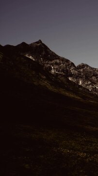 Mountains are seen under a dark sky in a remote area. The landscape shows a mix of shadow and light as evening falls. No people or structures are present.