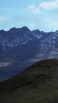 A view of large mountains with snow on the peaks. Green hills are in the foreground. The sky is clear and blue, indicating it is daytime.
