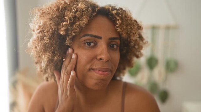 Woman touching cheek with hand, bra straps visible, slight smile and steady eye contact, natural window light and hanging plant in bedroom setting; skincare calm.