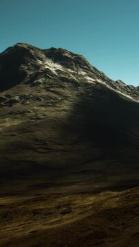 A mountain stands tall under a clear blue sky. The sun casts a long shadow on the ground. The landscape shows rocky terrain and grass below the mountain.