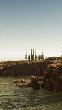 A rocky shoreline meets clear water. Green trees stand atop the cliffs. The scene shows a bright sky with few clouds. This place seems quiet and untouched by people.