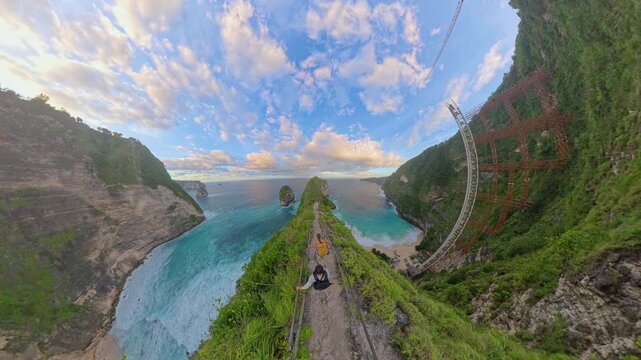 Panoramic View of Two Tourists Descending Narrow Ridge Toward Kelingking Beach on Nusa Penida Between Towering Coastal Cliffs