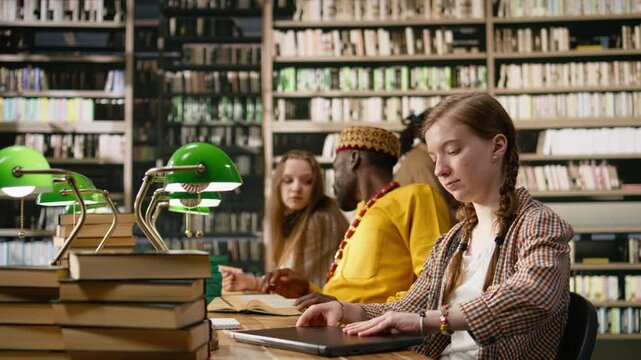 Young woman taking notes on laptop and studying beside open book at busy study table, peers using study materials and books, typing on keyboard near green lamps, for education technology.