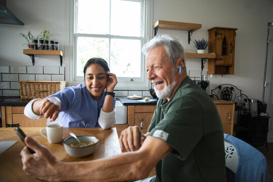 Senior man and caregiver using smartphone during breakfast in home kitchen