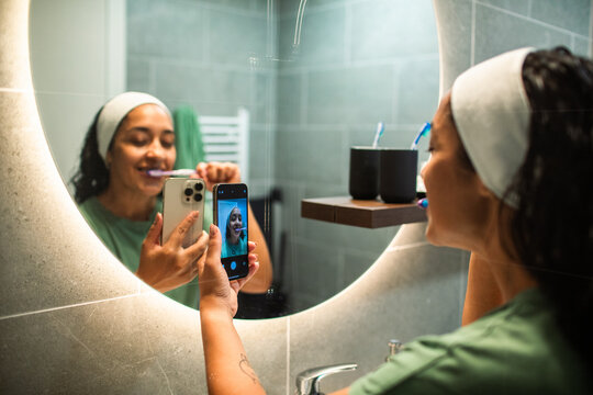 Woman brushing teeth and taking selfie in modern bathroom mirror