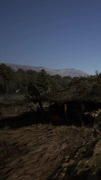 A cabin sits in the woods as morning light breaks over the landscape. Trees surround the cabin, and distant mountains are visible under a clear blue sky. The ground shows signs of wear.