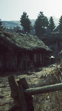 Several old houses with thatched roofs stand near a dirt path surrounded by trees. The sun sets, casting warm light on the scene. Grass grows along the path.