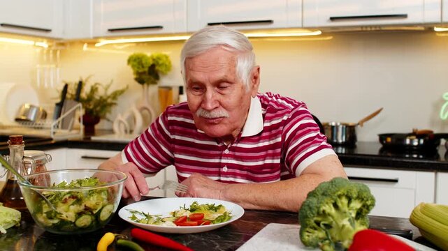 Senior old man smiling and eating fresh vegetable salad in kitchen at home for a healthy diet. Elderly grandfather enjoying weight loss nutrition and wellness meal with joyful satisfied mood indoors.