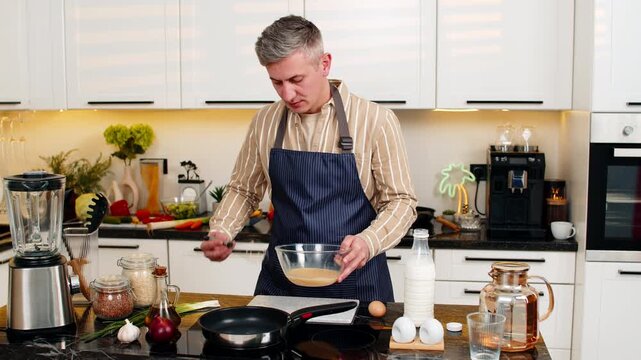 Middle-aged man pours omelet mixture into pan and fries healthy breakfast for diet meal in home kitchen with smile. Young adult guy enjoys cooking routine for nutrition weight loss and clean eating.
